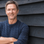 Homme souriant en pull bleu contre fond sombre.